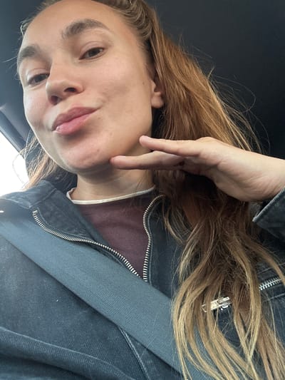 This is a close-up, waist-up, selfie-style photograph of a young woman with long, wavy, light brown hair. She is wearing a dark denim jacket over a maroon top with a cream-colored trim visible at the neckline. A dark gray seatbelt is diagonally crossing her chest. Her left hand is raised, with her index finger and thumb touching her chin. Her mouth is slightly pursed. The background is dark, suggesting an indoor setting or a vehicle. There is no visible indication of the city of Uva, Russia.