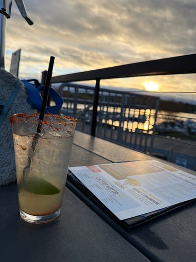 The image shows a drink on a table, likely at a restaurant or bar. A cocktail with a chili-salt rim and a lime wedge is visible, with a black straw inside the glass. Part of a menu can be seen next to the drink. The menu lists "Cactus Water" and "Hard Lemonade" as drink options. Outside the restaurant/bar, a glimpse of buildings is visible in the background, including a metal railing. The sky appears to be reflecting the sun, indicating it might be late afternoon or early evening. Location is Laconia, United States of America.