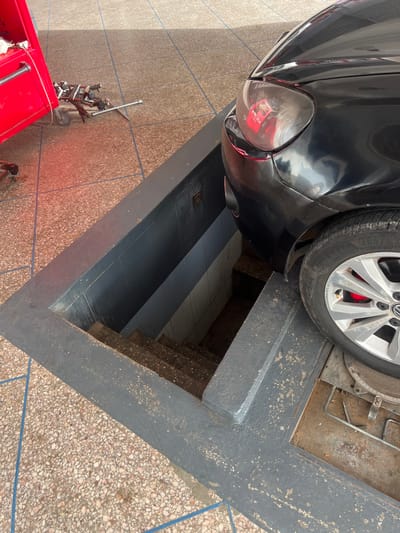 The image shows a black car undergoing maintenance. Its front left tire rests on the edge of a pit in a repair shop, exposing the wheel well. A red tool chest and tire alignment apparatus are visible on the textured tile floor to the left of the pit. The location is likely in Beni Mellal, Morocco, implied by the architectural style and materials used in the pit's construction and floor tiling. No people are visible. The scene depicts a car repair process.