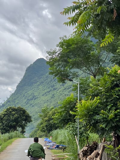 The image depicts a rural road in Lùng Tám Commune, Vietnam, with a person riding a motorcycle away from the camera. The road is paved and curves into the distance, flanked by lush green vegetation and tall trees. In the background, a large, verdant mountain dominates the skyline, shrouded in clouds. A street lamp is visible on the right side of the road, and piles of materials, possibly for construction or crafts, are stacked near the roadside. The sky is overcast with heavy clouds, suggesting a potentially rainy or humid day. The overall scene conveys a sense of natural beauty and remote rural life.