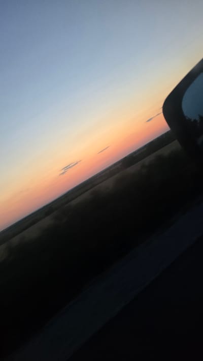 A tilted, close-up, outdoor shot captures a sky at dusk transitioning from a light blue at the top to a soft peach and pink gradient towards the horizon. Small, wispy clouds are scattered across the sky. Below the sky is a dark, silhouetted landscape, likely agricultural fields, that stretches across the frame. In the upper right corner, a portion of a dark, reflective car side mirror is visible, obscuring part of the sky and landscape. The scene is viewed from inside a vehicle, as suggested by the car mirror and the angle of the shot. The location is the city of Belotintsi, Bulgaria.