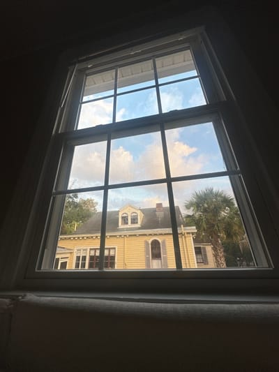 From inside a darkened room, a window frames a view of a bright, sunny day. A white-framed window with multiple panes looks out onto a two-story yellow house. A large palm tree stands near the building. The sky is blue with scattered clouds, some reflecting the colors of the setting sun. The angle of the window suggests it is a skylight.