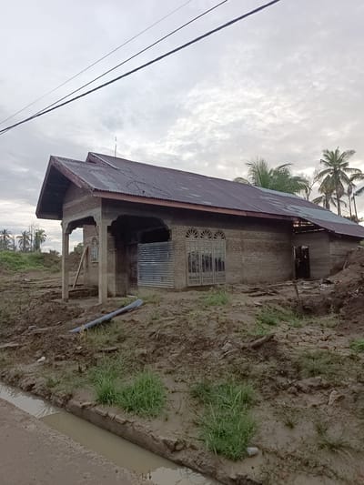 The image depicts an overcast day in Meunasah Raya, Indonesia. A partially constructed or dilapidated brick building with a corrugated metal roof stands on muddy ground. The building has a covered porch supported by concrete pillars, and the windows appear to be ornate. To the left of the building, a grey pipe is visible on the ground, and a small ditch filled with murky water runs in the foreground. Palm trees are scattered in the background, suggesting a tropical or subtropical climate. The overall scene conveys a sense of disrepair and unfinished construction within a rural or semi-rural setting.