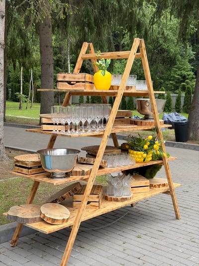 The image shows a rustic wooden bar set up outdoors, likely for an event. The bar is constructed from rough-hewn wood and features multiple shelves and tiered levels. On the shelves, various glassware is arranged, including wine glasses and tumblers, some with a textured pattern. A large, silver ice bucket sits prominently on one of the lower shelves. Decorative elements include a bright yellow faux lemon and a bunch of yellow artificial tulips with green leaves, adding a touch of color to the scene. A small section of moss-covered decor is also visible.

The setting appears to be a grassy area adjacent to a paved walkway, with mature trees and lush green foliage forming the backdrop. The overall impression is of a casual, outdoor gathering or celebration. The time of day seems to be daytime, and the weather appears to be overcast or mildly cloudy, given the even lighting and lack of harsh shadows. There are no people visible in the image. The location context suggests this might be in or near Pyatryshkaŭski Sel'ski Savet, Belarus, which is an area known for its natural beauty. The construction of the bar, with its exposed wood grain and rough finish, suggests a DIY or handcrafted aesthetic.