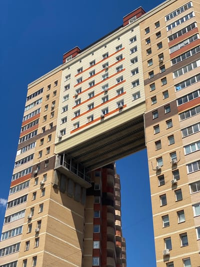 This is a photograph taken from a low angle, looking up at the exterior of a large, multi-story building against a clear, bright blue sky. The building's facade is predominantly light beige brick with alternating horizontal bands of darker brick and reddish-brown elements. Numerous windows are visible, arranged in regular patterns across the building. Some windows are open, revealing glimpses of interior spaces, while others are closed. Several air conditioning units are mounted on the exterior walls.
The building's architecture is complex and appears to be a single structure with different sections extending outwards and upwards, creating an unusual, almost cantilevered appearance in some areas. There is a distinct geometric design with sharp angles and protruding sections. A narrow, open-air staircase with stone steps can be seen ascending along one side of a protruding section of the building.
The lighting suggests it is daytime, with the sun casting shadows that highlight the building's architectural features. There are no people visible in the photograph. The overall impression is one of urban architecture and design, with a focus on the structural elements and the interplay of light and shadow on the building's surfaces. The clear blue sky indicates fair weather. The provided location context places this scene in Kotelniki, Russia. No text is visible in the image.
