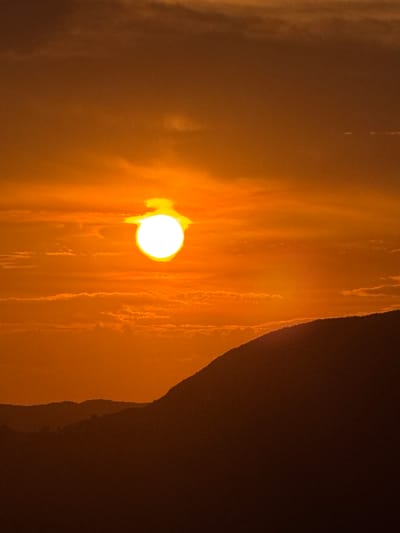 The media file displays a close-up view of a bright, circular sun or light source casting an orange glow. The light illuminates a textured, vertical surface that appears to be a waterfall or a rock face with flowing water, creating streaks and patterns. To the right, a dark, shadowed area suggests a cliff or a dense, unlit space. The overall impression is of a natural landscape at sunset or sunrise, with the warm light highlighting the dynamic movement of water against a dark backdrop. The location is identified as Čelobrdo, Montenegro. There is no visible text in the image.