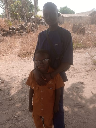 The scene, located in Gudige Sabo, Nigeria, depicts two individuals outdoors. A taller male, dressed in a dark blue short-sleeved top, dark pants, and a black shoulder bag strap, stands behind a shorter male, identified as a child. The taller individual has his right arm resting on the child's left shoulder and neck. Both individuals possess dark complexions and face the camera directly. The child wears an orange two-piece button-up outfit, oversized black-framed glasses, and a black beaded bracelet on his left wrist. The ground consists of light brown, dry earth with sparse dry vegetation. In the background, multiple bare trees are visible, along with several simple, single-story structures featuring light-colored roofs. Piles of cut logs and stones are present in the mid-background. The environment is illuminated by bright daylight.