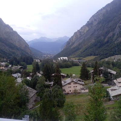 This image shows a panoramic view of the town of Courmayeur, Italy, nestled in a valley between dramatic, tree-covered mountains. The foreground is dominated by the roofs of traditional stone-and-slate houses, characteristic of alpine architecture. Dense evergreen trees and some deciduous trees are scattered throughout the scene, with some partially obscuring the view of the houses.

In the midground, several larger buildings are visible, including a notable white structure with a distinctive, modern architectural design that appears to be a public building or resort. This structure is surrounded by green lawns and more trees. Further into the valley, the town of Courmayeur sprawls out, with a denser concentration of buildings visible on the slopes and in the distance, leading towards more distant, hazy mountain ranges. The sky is overcast with a pale, diffused light, suggesting either early morning, late afternoon, or a cloudy day. There are no visible people or animals in the frame. The overall impression is one of a serene, picturesque mountain town on a subdued day.