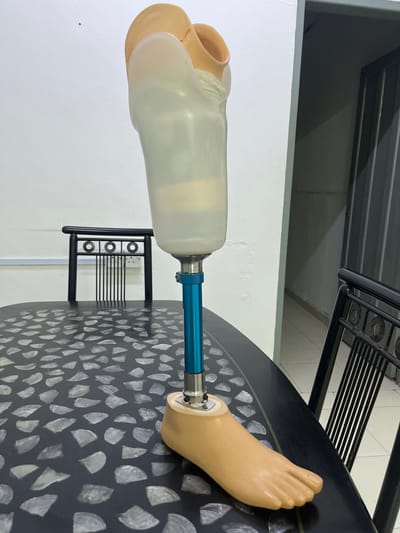A prosthetic leg is on a black table with an irregular pattern. The table is next to a white wall. A metal rack with a black frame is visible in the background. The location is likely in Ipoh, Malaysia.