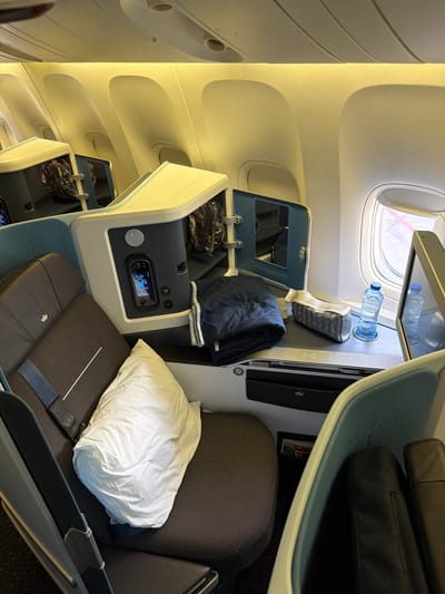 The image shows a view of business class airplane seats, likely inside a KLM Royal Dutch Airlines aircraft, as indicated by the crown logo on the headrest of the seat in the foreground. The seats are in a forward-facing, staggered configuration, with a privacy shell surrounding each seat. The seat in the foreground is dark brown with a light-colored pillow placed on it. A blue seatbelt is visible.
The immediate area around the seat includes an open compartment with items inside, a folded navy blue blanket, a small toiletries pouch, and two bottles of water. A digital display is embedded in the console. A window with a view of the tarmac is to the right, with another airplane seat visible beyond it.
The lighting is warm and appears to be from the overhead cabin lights. The overall environment suggests a parked aircraft ready for boarding or during a flight. Given that KLM's main hub is Amsterdam Airport Schiphol (AMS), it is highly probable that this image was taken at Schiphol, Netherlands. However, the image itself does not contain explicit details to confirm the specific location within Schiphol, such as gate numbers, terminal signage, or distinctive architectural features outside the aircraft.