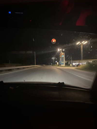 The image captures a nighttime scene from what appears to be the interior of a vehicle, looking out through a windshield or window. The perspective is slightly angled, suggesting movement.
The main subject is the road ahead, illuminated by streetlights and the lights of a building that resembles a gas station or convenience store. The building has prominent signage, including what looks like a "Shell" logo and price displays. Multiple lights are visible on poles and on the building itself, casting a glow on the wet asphalt of the road, indicating either recent rain or dew.
To the left of the road, there are dark areas with hints of red light, possibly from interior lights of the vehicle or reflections. A circular, red and white object, possibly a decorative light or a warning signal, is visible in the lower left quadrant. Further back, more lights suggest the presence of other structures or vehicles.
The overall atmosphere is dark and nocturnal, with artificial lights being the primary source of illumination. There are no people clearly visible, but the scene implies human activity through the presence of the road and the commercial establishment. The time of day is unequivocally night. There are no explicit weather cues beyond the potential wetness of the road.
The location context of Yasen, Bulgaria, provides a geographical anchor, but no specific visual cues within the image directly confirm this location. The visible text, "Shell," is a brand name common globally. The scene evokes a sense of travel or a stop during a nighttime journey.