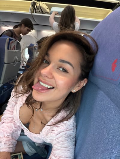 A young woman is taking a selfie on an airplane. She is seated and leaning back against the headrest, sticking out her tongue and smiling at the camera. She has brown hair and is wearing a pink top with a low neckline and a double-strand necklace. Other passengers can be seen in the background, along with airline seats and overhead compartments. The image does not provide enough detail to determine the exact location as the Shunyi District in China.