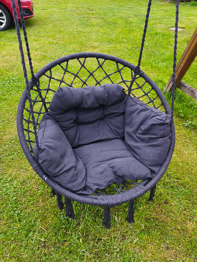 An empty, dark grey fabric cushion sits inside a black macrame hammock chair. The chair hangs from thick black ropes attached to a wooden support structure, partially visible on the right. The chair rests on a well-maintained green lawn. In the background, a portion of a red car is visible on the left side of the frame. The overall scene suggests an outdoor setting, possibly a backyard or park. The location is Brodi, Latvia.