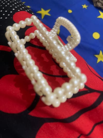 A close-up shot captures a pearl hair claw, an accessory that appears to be made of plastic beads, resting on a patterned fabric. The fabric beneath features a red background with black and white polka dots on the left side and a bold red pattern on the right. The hair claw itself is white and appears to be intricately crafted from small, round beads. The background is a blue fabric adorned with yellow stars and circles, suggesting a playful or decorative design. The image is taken from a top-down perspective, focusing on the hair claw and its immediate surroundings. There are no people or discernible actions depicted. The scene is static and focuses solely on the object and its placement on the fabric. No specific location in Anggaswangi, Indonesia is identifiable from the visual information provided.