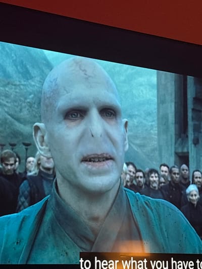 A close-up, tilted shot shows Voldemort, from the "Harry Potter" series, with a pale face, no nose, and narrow eyes. Behind him, a line of Death Eaters is visible. A blurred, vertical caption on the right reads "to hear what you have to". The image is framed by a black border, likely a TV screen, with a hint of orange wall on the left.