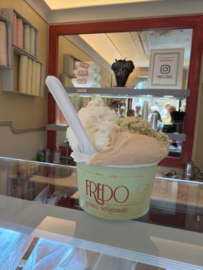 In Pienza, Italy, this close-up shot captures a vibrant gelato cup displayed at eye level in a glass case. The cup, a pale green with "FREDO gelato artigianale" emblazoned in red lettering, is brimming with three distinct scoops of gelato: a pale cream color, a light peachy hue, and a textured, light green flavor. A white plastic spoon is inserted into the gelato, suggesting immediate consumption.

Behind the gelato, a red-framed mirror reflects the interior of what appears to be a shop. Shelves filled with colorful, cylindrical paper cups are visible on the left. The mirror also shows a display with stacked white boxes with red labels, a black sign shaped like an affogato with "AFFOGATO AL CAFFÈ" written on it, and a framed Instagram post with a green border and a white Instagram logo. Blurred figures of customers and staff are visible in the reflection, engaged in shopping activities. The background is a warm, earthy yellow, and the overall lighting is bright and inviting. The glass counter in the foreground, covered with protective plastic, hints at a retail environment.