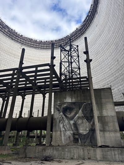The photograph captures an interior view of the cooling tower at the Chernobyl Nuclear Power Plant in Pripyat, Ukraine. The massive, curved concrete structure of the cooling tower dominates the upper portion of the image, with scaffolding visible along its rim. Inside, a complex network of decaying concrete pillars and metal supports creates a skeletal framework. A prominent mural of a pensive human face, rendered in black and white, is painted on a concrete wall within the structure, serving as a stark artistic contrast to the industrial decay. Large, dark pipes, likely part of the plant's infrastructure, run horizontally through the lower section of the scene. Overgrown vegetation and debris litter the ground at the base of the structure, indicating abandonment and the passage of time. The sky above, visible through the top of the tower, is partly cloudy.