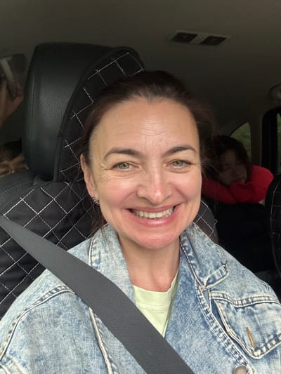 The image shows a woman in the front seat of a car, smiling at the camera. She is wearing a denim jacket over a light green t-shirt and her seatbelt is visible. In the background, two children are seated in the back of the car. The child on the right is leaning on a red pillow and appears to be asleep. The environment is indoors, inside a vehicle, with a dark interior. The image does not provide any specific location details within the city of Kulmas, Russia.