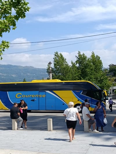 A large blue and yellow tour bus, labeled "Courense," is parked on a street. Several people are gathered near the bus. In the foreground, a woman in a white t-shirt and black skirt walks away from the camera. To her right, a man in a white polo shirt and gray shorts sits on a concrete bollard, talking to a woman in a blue dress who stands with a white plastic bag. Further left, a man in a black shirt and gray pants sits on another concrete bollard, facing a woman in a black dress who holds a pink water bottle. The bus has "AVIS" written on its side. Behind the bus, lush green trees and rolling, mountainous terrain with scattered buildings suggest a rural or semi-rural setting in Terras de Bouro, Portugal. A stone monument with a cross is visible behind the bus. The sky is bright blue with scattered white clouds, indicating it is a sunny day. The scene appears to be a stop for the bus passengers.