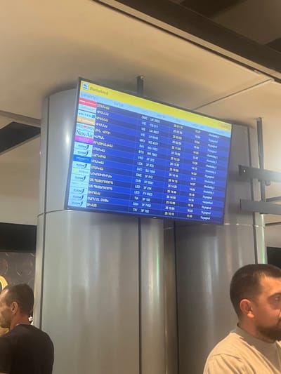 The image shows a flight information display board at an airport. The board displays flight numbers, destinations, and departure/arrival times in Armenian and English. The scene is likely indoors, given the lighting and the presence of a large digital screen. A few people are visible, partially out of focus, with their attention directed towards the display. One person in the foreground is looking down and to the right, while another is partially visible in the background, looking towards the screen. The lighting suggests it might be daytime, though the indoor setting makes it difficult to determine the exact time of day or weather conditions. The presence of an airport flight board indicates the location is an airport terminal. Text on the board is predominantly in Armenian, with some English flight codes and airline names. The overall impression is one of transit and travel.