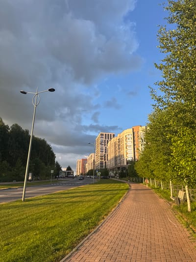 This image depicts a street scene in Koltsovo, Russia, likely during the late afternoon or early evening, as indicated by the warm, golden light casting long shadows across the meticulously paved walkway and the lush green grass. The scene is dominated by a row of modern, multi-story apartment buildings that are predominantly beige and light brown, with numerous windows reflecting the sky. The buildings appear residential and well-maintained.
To the left of the buildings, a vibrant green canopy of trees, possibly birch trees due to their leaf shape and density, overhangs the scene. The sky above is a mix of bright blue and dramatic, dark gray clouds, suggesting a change in weather or the lingering presence of a storm. A modern street lamp with a unique, sculptural design stands tall in the foreground, its metallic structure catching the light.
The paved walkway curves alongside the road, bordered by well-kept grass. The road itself is visible, with a few cars parked or driving along it, and there are pedestrian crossings and street signs, including a blue sign with a yellow pedestrian symbol. A bus stop shelter is also visible. While there are a few indistinct figures on the walkway and near the bus stop, no specific individuals or activities are clearly discernible. The overall impression is one of a modern, developing urban or suburban environment bathed in the golden light of a late day, under a dynamic sky.