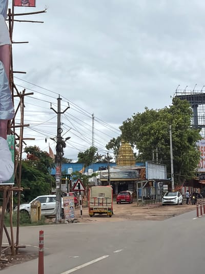The image depicts a street scene in Shamshabad, India, on an overcast day. A white car is parked on the left, partially obscured by wooden scaffolding and a large banner. In the background, a golden-domed temple structure is visible amidst trees. Various shops and buildings line the street, with a traffic signal and power lines overhead. An auto-rickshaw is parked near a building, and a red car is seen further down the road. A white hatchback is parked on the right, and a person is standing near it, alongside a motorcycle. Another person is visible on a motorcycle in the distance. A large billboard structure is present in the upper right corner of the image. The road surface is asphalt, with painted lane markings. Red and white traffic cones are placed on the side of the road.