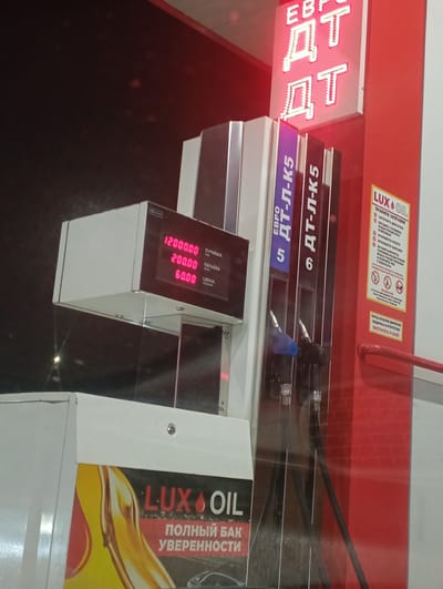 The image shows a gas station, likely in Enem, Russia, based on the Cyrillic lettering. The gas pump displays "EURO DT-L-K5" and "DT-L-K5" for the fuel types on pump 5 and 6 respectively. The pump's display shows "12000.00," "200.00" and "60.00" for "SUMMA" (amount), "OBYEM" (volume), and "TSENA" (price). A "LUXOIL" advertisement is affixed to the pump. A sign provides refueling instructions in Russian. The environment appears to be dimly lit, suggesting it may be nighttime.