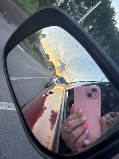 This is a mirror image taken from inside a vehicle, showing a reflection of the road and sky. The vehicle appears to be in motion, as indicated by the motion blur in the reflection of the road. The scene is set in the village of Akberdino, Russia. In the foreground of the reflection, a pink smartphone is being held by a person's hand, which has white and neon pink nail polish. The person's face is also visible in the reflection, looking towards the phone. The background of the reflection shows a road with traffic, trees, and a bright sunset sky. The car's red body is visible in the lower part of the frame, reflecting the sky and trees.