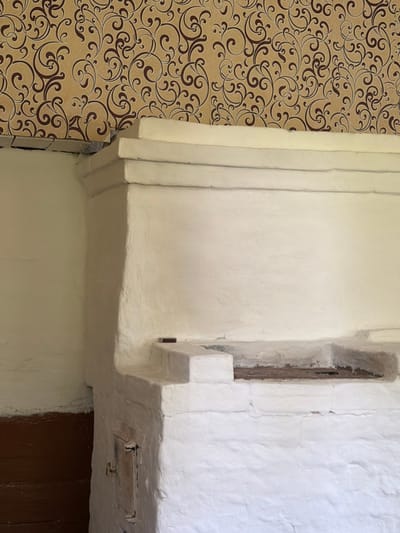 The image shows the interior of a rustic room. The dominant feature is a large, white, plastered stove, likely a traditional Russian stove (pech). The stove has a rough texture, suggesting it is made of brick or clay and has been whitewashed. There are visible cracks and imperfections in the plaster. The stove has a rectangular opening near the bottom, likely a firebox with a metal door, and a horizontal shelf-like structure above it, possibly for heating or cooking.

The wall above the stove is covered with wallpaper featuring an intricate, swirling pattern in brown and beige. The wallpaper appears to be older, with some slight damage visible near the corner where it meets the stove.

To the left of the stove, a portion of the wall is visible, also plastered and painted white, with a darker wooden paneling at the bottom, possibly wainscoting.

The overall impression is one of a simple, functional living space, likely in a rural or older dwelling. The scene is static and does not contain any people or immediate action. The lighting suggests an indoor environment, possibly with natural light coming from an unseen source. The specific location in Kameshki, Russia, implies a context of traditional Russian architecture and domestic life.