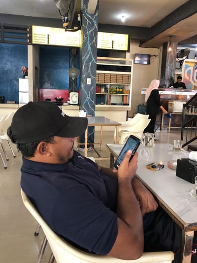 The image captures an indoor scene, likely a restaurant or cafe, in Keudeteupinpunti, Indonesia. In the foreground, a man wearing a navy blue polo shirt and a black baseball cap is seated at a table, engrossed in his smartphone. His right hand holds the phone, displaying a screen with text and an image. On the table in front of him are a pack of "CHIEF" cigarettes with one lit, an ashtray, and a glass of water.
In the middle ground, a woman wearing a light pink hijab and a black outfit walks away from the camera, towards a counter area where a man in a black shirt stands. Behind them, shelves are stocked with various items, including fruits and boxes.
The background reveals the restaurant's decor, featuring dark blue walls, a ceiling fan, and a bright yellow menu board with Indonesian text, listing food items. Another menu board is visible on the right side of the image, also displaying text. The overall environment suggests a casual dining establishment.