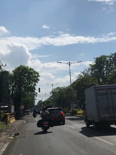 This is a street-level, full-shot image taken outdoors in Buduran, Indonesia, on a sunny day with a bright blue sky and scattered clouds. The perspective is from a moving vehicle, looking forward. The foreground is dominated by a paved road with white lane markings. In the middle ground, traffic includes a rider on a scooter carrying a large black bag, a red car, and a large white truck. Several other vehicles and riders are visible further down the road. The background features lush green trees lining the street and utility poles with power lines. A traffic light is visible on the left side of the road. The overall scene depicts a typical street scene with moderate traffic on a clear day.