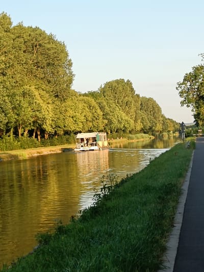 An outdoor, eye-level, vertical shot shows a canal with a houseboat floating on it, on a sunny day. The canal water is yellowish-brown, with reflections of the trees and sky. The left side of the canal is lined with lush green grass and dense trees, their leaves illuminated by the sun. On the right side, a paved path runs parallel to the canal, bordered by grass. The sky is a clear, pale blue. The houseboat is white and yellow, with a covered section. Several people appear to be seated on the bank of the canal to the left of the houseboat. The scene is likely a scenic route within or near Haacht, Belgium, given the prevalence of canals and tree-lined waterways in the region.