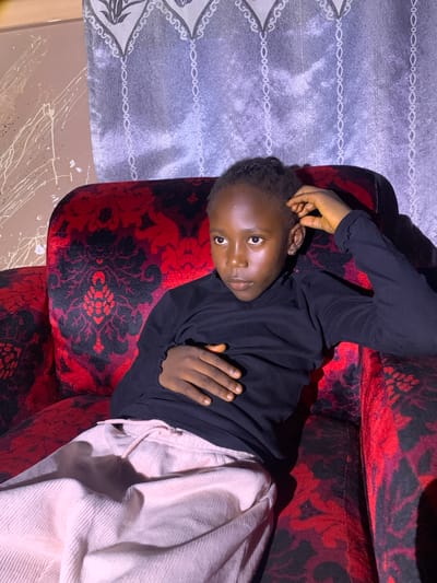 A young person with dark skin, wearing a black long-sleeved shirt and light pink pants, is lying on a patterned red and black couch. The person is lying on their right side, facing the camera with their head propped up by their left hand. Their right arm is resting on their stomach, with their hand flat. The couch has a floral damask pattern. To the left of the couch, there is a grey curtain with a white embroidered pattern. The lighting appears to be artificial, suggesting it could be indoors, possibly at night or in a dimly lit room. The overall mood seems relaxed, with the subject looking directly at the camera. There is no visible text in the image. The location context is Russo, Nigeria.