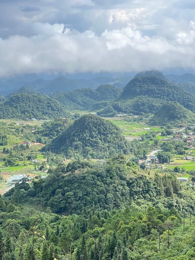 The image captures a panoramic view of a lush, mountainous landscape, characteristic of the Quản Bạ Commune in Vietnam. The scene is dominated by numerous rolling hills covered in dense green vegetation, with some appearing as conical formations. In the valleys between the hills, there are patches of cultivated land, likely rice paddies, showcasing vibrant green hues. Scattered throughout the landscape are small villages with houses featuring red-tiled roofs, interspersed with trees and roads. A winding road is visible, leading through one of the valleys. The sky above is partly cloudy, with large, soft white clouds obscuring parts of the distant mountain ranges. The overall impression is one of a serene and verdant rural environment. There are no people or animals clearly visible in the image. The foreground is filled with trees, some with bare branches, creating a textured layer to the overall composition.