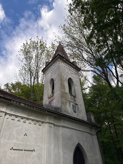 The image captures the tower of the Chapel of the Assumption of Mary in Častá, Slovakia, from a low-angle perspective. The tower is made of a light-colored, possibly stone material, and features a small, tiled spire at its peak. Two arched openings are visible on the tower, with decorative elements surrounding them. Above the upper arch is a small, carved emblem. The side of the chapel has decorative crosses. The surrounding environment includes trees and a blue sky with clouds. Branches and leaves of nearby trees frame the building. The sky is partially visible through the trees.