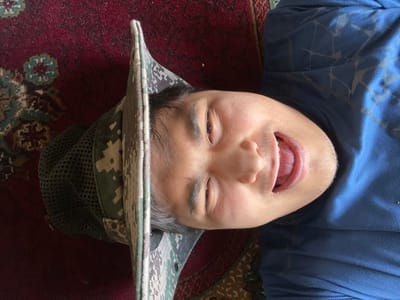 A single individual is observed from an overhead perspective, lying on a patterned red rug within the city of Mirzabashichek, Uzbekistan. The subject, a male with medium complexion, dark hair, and dark eyes, wears a blue t-shirt with a subtle geometric pattern and a digital camouflage boonie hat featuring a mesh crown. He is positioned horizontally, with his head turned towards the camera, mouth open, tongue partially visible, displaying a wide smile. The underlying rug is crimson-toned with traditional floral and geometric designs in blue-green and light beige.