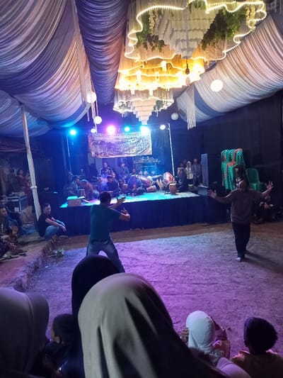 An evening view captures an outdoor or semi-outdoor cultural performance taking place under a large, draped tent or canopy in Kecamatan Cimaung, Indonesia. The ceiling of the tent is adorned with flowing white fabric, intricate decorative lighting resembling a chandelier with warm white lights and artificial foliage, and hanging white lanterns, creating a festive atmosphere.

At the back of the scene, a raised stage is brightly illuminated by blue, purple, and pink stage lights. Several musicians are seated on the stage, playing traditional Indonesian instruments, possibly gamelan or similar percussion. A large banner hangs behind them, displaying the text "LINGKUNG SENI 'PUTRA JAYA'" (Art Environment 'Putra Jaya'), with smaller text underneath indicating "MINGGU, 10 NOVEMBER 2024" (Sunday, 10 November 2024), along with other less discernible information. Stacked green plastic chairs are visible on the right side of the stage.

In the foreground, in front of the stage, two men are performing on the dirt ground, which is cast in purple light. One man, in a green shirt and jeans, is captured mid-movement, appearing to dance or execute a martial arts pose with his arms raised. Another man, to his right, is also performing with outstretched arms.

The audience, including individuals wearing headscarves and some children, is seated on the ground in the foreground and around the performance area, observing the event. The overall scene suggests a community gathering or festival celebrating local arts and culture.