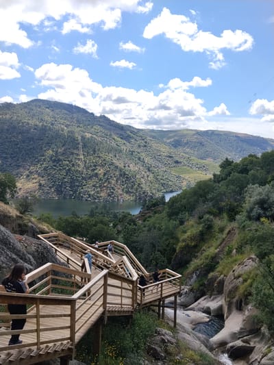 The image captures the wooden walkways of Passadiços do Tua, located in Alijó, Portugal. People are walking and standing on the walkways, which are constructed along a rocky, vegetated slope. Below, there is a stream with small waterfalls. In the background, a large river surrounded by forested hills is visible under a sky with scattered clouds. Overhead power lines stretch across the landscape in the distance.