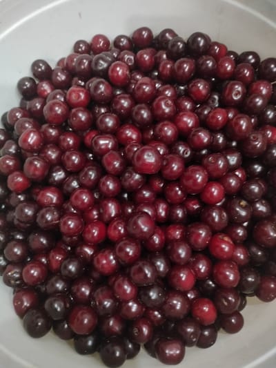 The image shows a close-up, overhead view of a white bowl filled with dark red cherries. The cherries are round and have a glossy sheen, with some appearing slightly wrinkled. They are piled high in the bowl, obscuring much of its interior. The lighting is somewhat dim, casting subtle shadows on the cherries. The scene is static, with no visible people or action. This image does not depict a location in the city of Kalach, Russia.