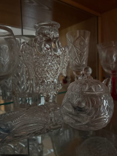 The image displays a collection of clear glass decorative items, including a lidded sugar bowl with ornate etching, a glass figurine that resembles a standing owl with intricate detailing, and several glasses with cut glass patterns. The objects are arranged on glass shelves within a wooden cabinet. The lighting is subdued, highlighting the translucent quality of the glass and creating reflections. The background consists of the dark wood of the cabinet and appears to be an indoor setting. This image is not specific to Tutaev, Russia, nor does it contain any information that would link it to that location.