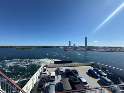 The image shows a ferry deck laden with cars, sailing on a bright, sunny day. The sky is a clear, deep blue with a prominent sun flare streaking across the upper right quadrant. In the distance, the city of Karlskrona, Sweden, is visible, characterized by industrial structures including tall, slender towers and lower buildings lining the waterfront. The water is a dark blue, with the ferry creating a white, foamy wake. Several white seagulls are scattered across the water. The ferry deck is organized with rows of parked vehicles, including SUVs, vans, and sedans. The railing of the ferry is visible in the foreground. The overall scene depicts a journey with industrial coastal scenery.