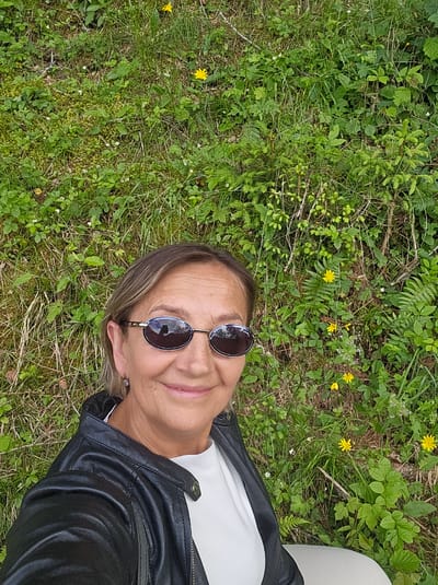 A woman, appearing to be of middle age, is pictured in a selfie taken outdoors. She is wearing a black leather jacket over a white top and sunglasses. She has short, light brown hair and is smiling. The background consists of lush green vegetation, including grass, small plants, and yellow wildflowers. The image does not provide any information indicating the location is Pontagna, Italy.