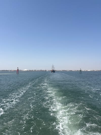 The image shows a view from a boat looking out at the sea towards a coastal city. The foreground is dominated by the foamy wake left by the boat. In the middle ground, a tall, dark sailing ship with multiple masts is visible, moving away from the viewer. To the left of the sailing ship, a red and white buoy stands in the water. To the right, a green and white buoy is also present. The background features a sprawling coastline with numerous buildings, suggesting a populated area. The sky above is a clear, bright blue, indicating a sunny day. The location is identified as the sea near Houmt Souk, Tunisia, with the coastline possibly representing the city's edge. There are no people clearly visible in the image.