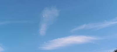 This image depicts a bright blue sky with scattered wispy clouds and numerous small dark specks, likely birds or insects, dispersed throughout. A thin crescent moon is visible in the upper center of the frame, partially obscured by a tall, elongated cloud formation. Another broader, fainter cloud stretches horizontally across the lower portion of the sky. The scene appears to be an outdoor shot taken in the city of Merchalovo, Russia, possibly during daylight or late afternoon hours, given the bright sky. There are no visible people or man-made structures within the frame.