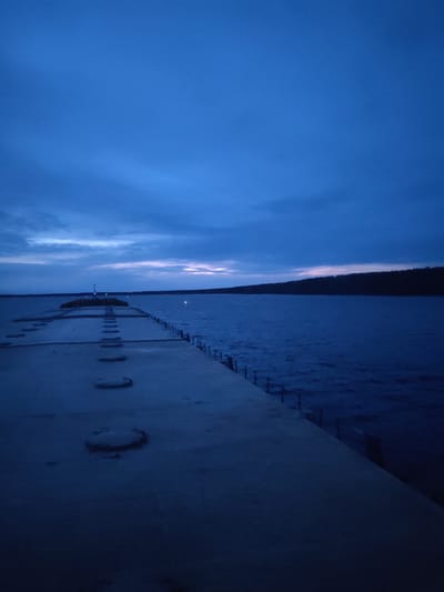 A low-angle, long-exposure photograph captures a concrete pier extending into a body of water, likely taken at twilight or dawn. The pier is the primary subject, and it stretches out from the foreground towards a distant structure with a light source, possibly a lighthouse or marker. The pier has several circular indentations along its surface. A metal railing lines the side of the pier closest to the water. The water appears choppy with small waves. On the opposite side of the water is a dark shoreline of trees. The sky is mostly covered in dark blue clouds. This image appears to be taken in Topornya, Russia.