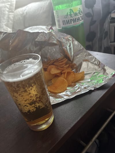 A close-up, eye-level shot captures a glass of beer with a frothy head and bubbles, placed on a dark, textured surface. Next to the glass, a crinkled silver bag contains a pile of ridged potato chips. In the background, out of focus, a green bottle with a label featuring mountains and Cyrillic text, along with a blurred white pillow or blanket, are visible. The scene is dimly lit, suggesting an indoor setting. The specific location in the city of Karadjovo, Bulgaria, is not discernible from the visual information provided in the image.
