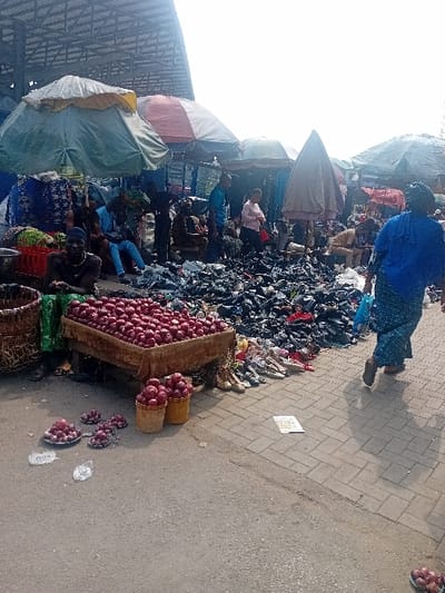 The image portrays an active outdoor market scene situated in Ebute-Metta, Nigeria, under bright daylight. The ground surface consists of dark asphalt in the immediate foreground, transitioning to grey brick paving on the right side of the frame.
In the foreground, a wooden stall is laden with a substantial quantity of red onions. To its right, two yellow buckets are also filled with red onions, and several smaller arrangements of onions are displayed on white plates directly on the asphalt, accompanied by white price tags. A dark-skinned individual wearing a dark top is seated on the ground behind the main onion stall, facing the viewer.
The mid-ground is occupied by multiple market stalls shaded by various umbrellas, including a prominent green umbrella, a red-and-white striped umbrella, and several darker-toned umbrellas. Beneath the green umbrella, another person is seated. A large, dispersed pile of black footwear, primarily shoes and sandals, occupies a significant area of the paved ground, with a linear display of lighter-colored footwear adjacent to it. Further back, several individuals, both standing and seated, are visible among the stalls, including a person in a light pink top and another in a light blue top. On the far right, a female figure, viewed from behind, walks from right to left across the brick paving. She wears a blue head covering, a patterned blue top, a long patterned skirt, and carries a blue plastic bag in her left hand.
The background reveals the exposed metal truss structure of a large building's roof, indicating a permanent market facility overhead. The scene depicts a bustling commercial environment with multiple vendors and market-goers.