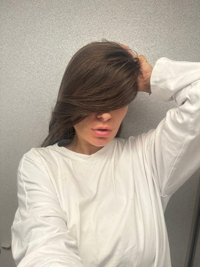 A person with dark brown hair covering their forehead lies on a textured, light gray surface. They wear a long-sleeved white shirt. Their hand rests on their head. The face is partially obscured by hair, revealing the mouth, nose, and part of the cheek. The lighting appears soft and diffused. Several stains are present on the shirt. The image is rotated counter-clockwise.