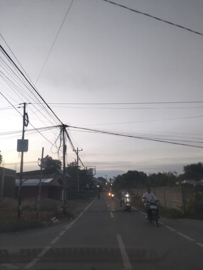 This media file depicts a street scene in Meunasah Papeun, Indonesia, during the late afternoon or early evening. The sky is overcast, suggesting cloudy weather. The foreground is dominated by a road with a dashed white line marking the lane. Several motorcycles are visible on the road. In the middle ground, two men are riding motorcycles, one with a white shirt and the other with a dark shirt. To the left of the road, there are buildings with corrugated metal roofs and a utility pole with a dense network of wires. The right side of the road features a fence and some vegetation. The overall impression is that of a quiet, everyday scene in a semi-urban or rural Indonesian setting, with the approaching dusk casting a subdued light. There is no visible text in the image.