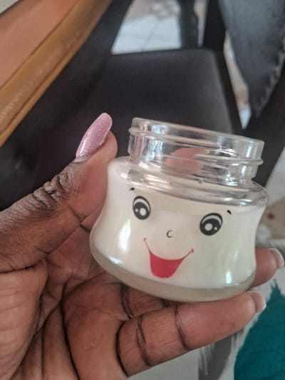 An individual's dark-skinned hand is shown holding a small, clear glass jar in an indoor setting within Muthambi ward, Kenya. The hand displays an elongated, pointed, glittery pink artificial nail on the index finger. The jar, which is approximately half-filled with a translucent, off-white substance, features a cartoon-style face printed on its front, consisting of black circular eyes, small black eyebrows, a black 'C' shape representing a nose, and a wide red smiling mouth with a visible tongue. The jar's top has a threaded rim. The background is blurred, revealing segments of what appears to be a light brown wooden surface, a dark piece of furniture (possibly a chair), and a patterned fabric in teal and white.